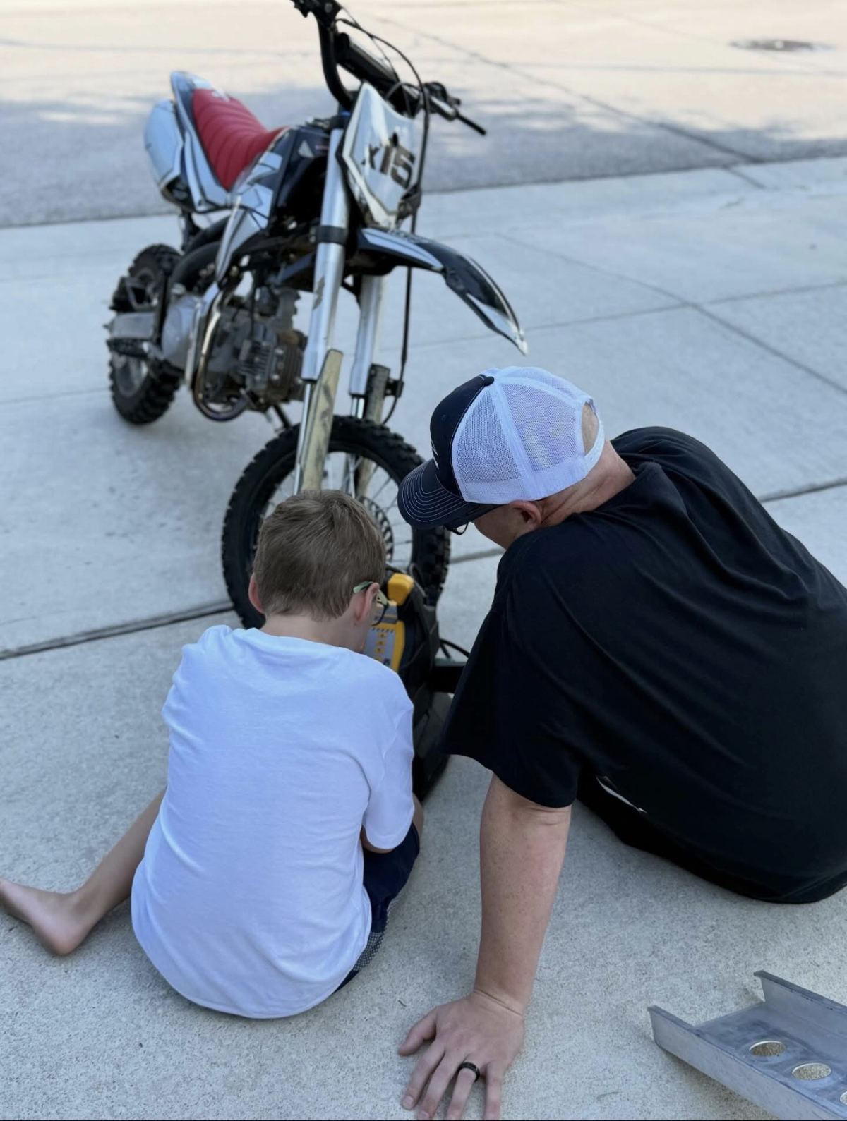 A father and son sitting on the driveway together, working on a dirt bike